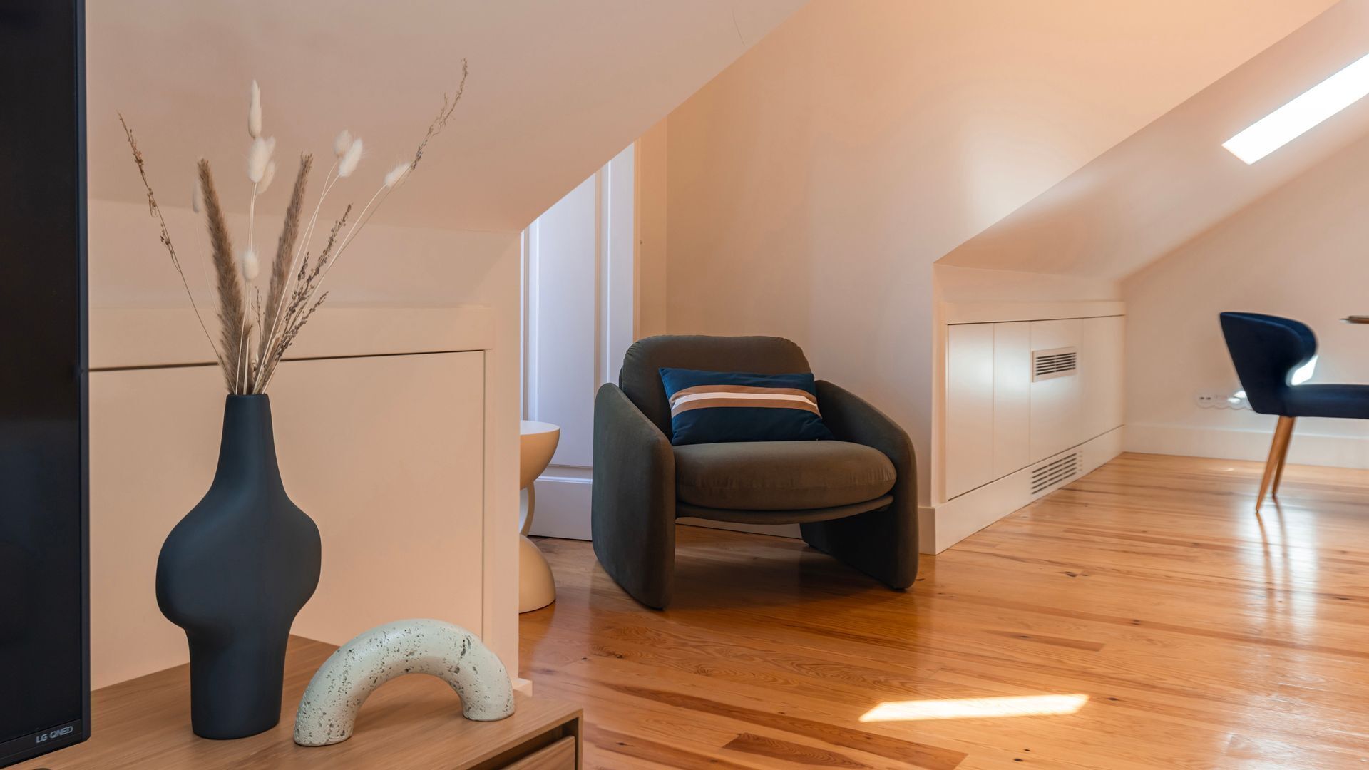 Ein modernes Interieur mit einer gebogenen Bank, einer Vase mit Trockenpflanzen und einem gewölbten Holzelement auf Holzdielen.