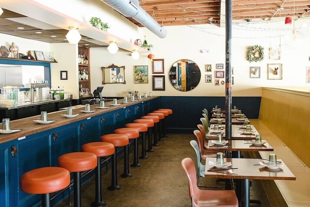 Un restaurante con una barra larga, taburetes naranja y mesas dispuestas para comer.