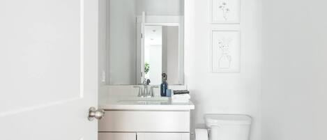 Un baño blanco con lavamanos, espejo e inodoro. Hay cuadros enmarcados en la pared.
