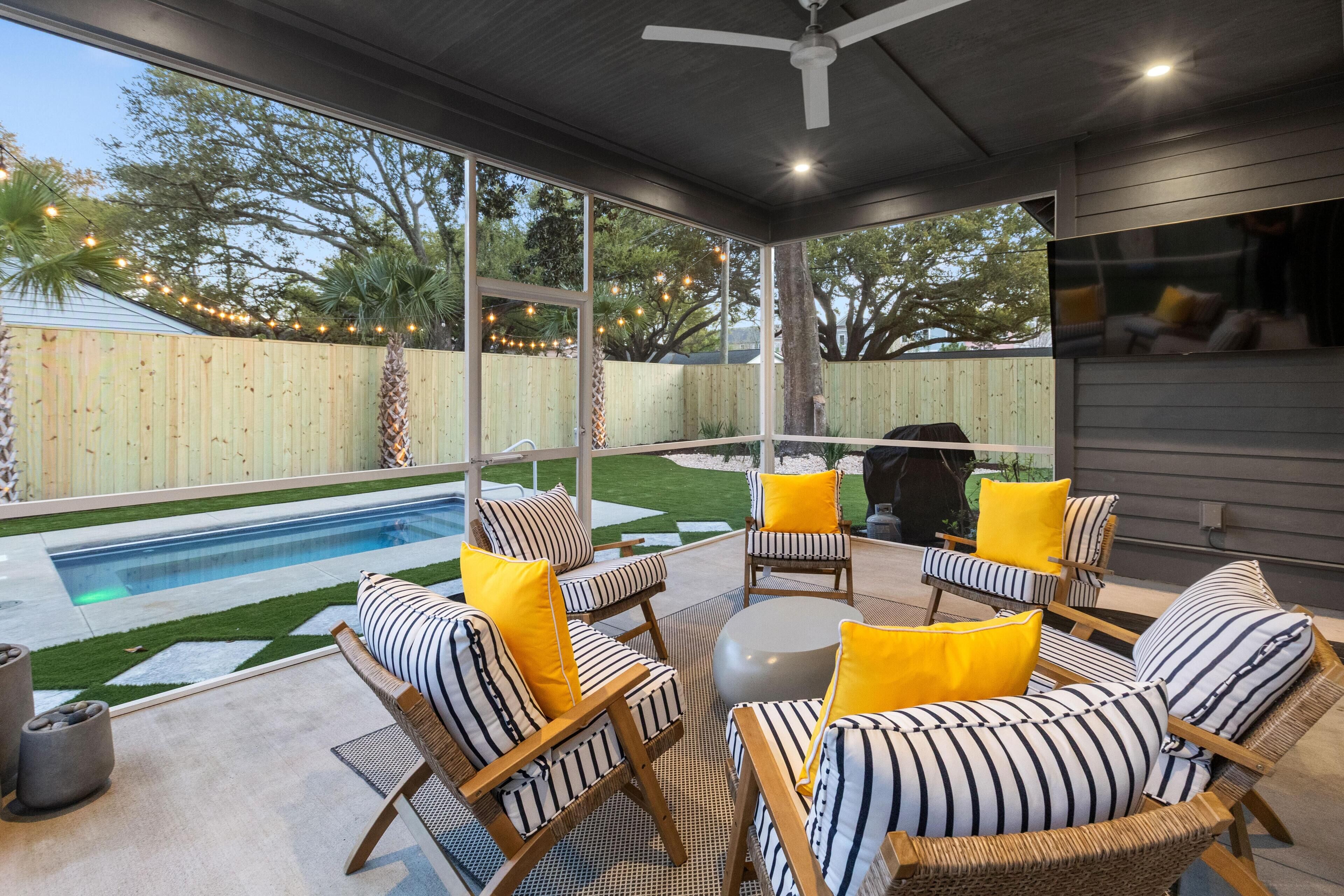A covered outdoor seating area with wicker chairs and cushions, a pool, and a view of a backyard with a wooden fence and trees.