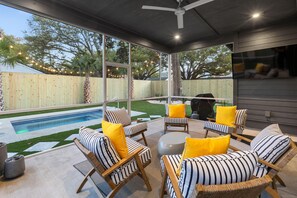 A covered outdoor seating area with wicker chairs and cushions, a pool, and a view of a backyard with a wooden fence and trees. - Music Manor-3-king Beds-pool-mins To Beach & DT (Mount Pleasant)