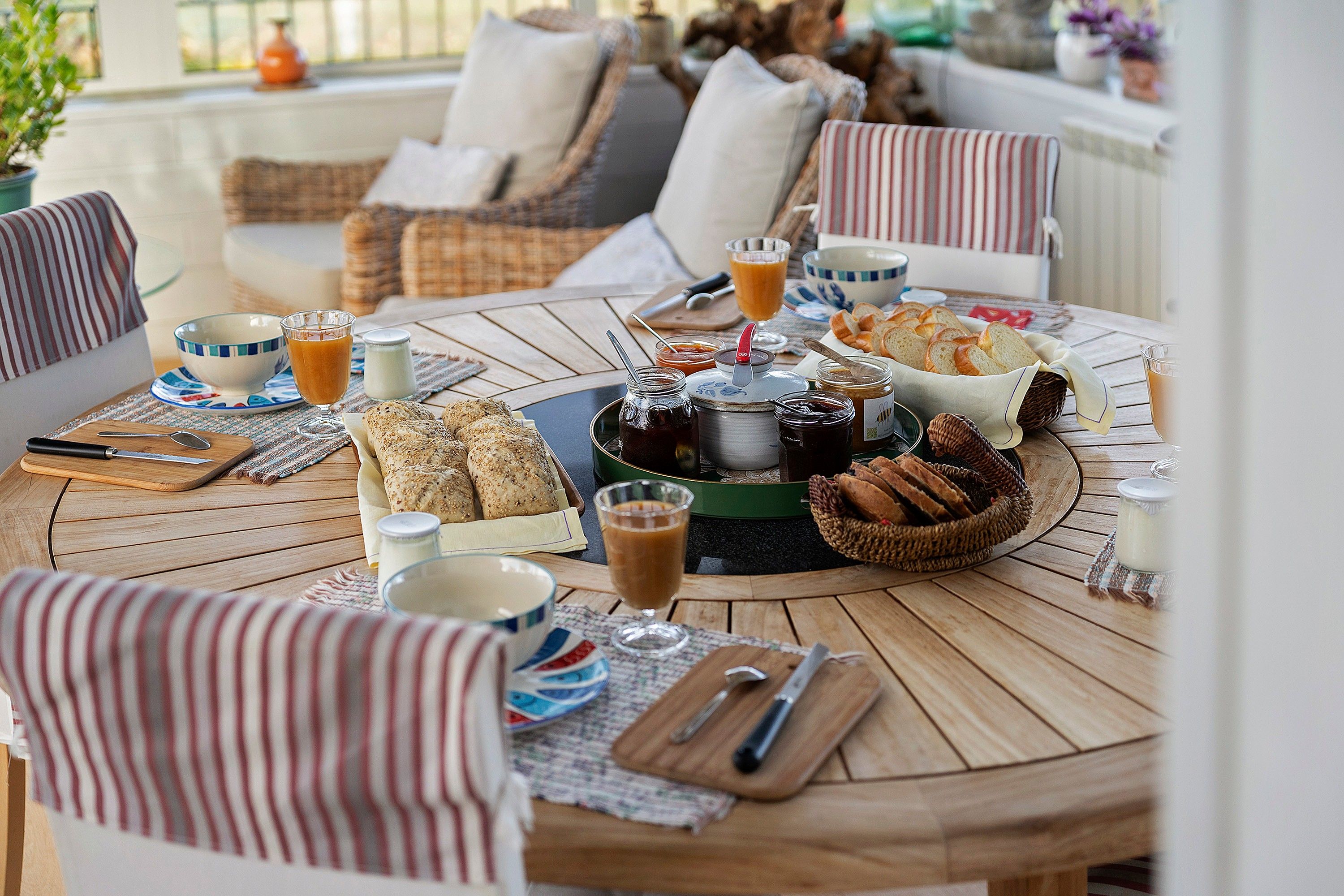 Een ronde houten tafel met een ontbijtsetting, inclusief brood, boter, jam en drankjes.