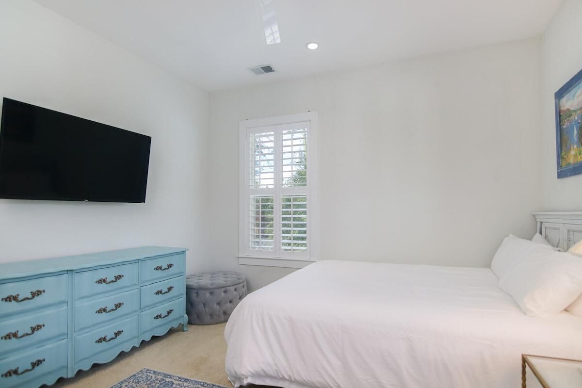 Ein Schlafzimmer mit einem Bett, einem an der Wand montierten Fernseher, einer Kommode und einem Fenster mit Jalousien.