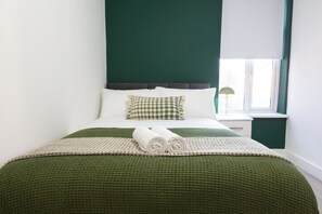 Ein ordentlich gemachtes Bett mit einer grünen Decke, zwei aufgerollten Handtüchern, einem karierten Kopfkissen und einem Nachttisch mit einer Lampe.