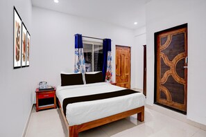 A bedroom with a bed, a nightstand, a window with curtains, a wooden door, and a wall-mounted artwork. - Family Rooms With King bed Ravai (Calangute)