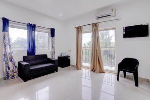 A modern living room with a black sofa, a small black table, a TV mounted on the wall, and large windows with curtains. - Family Rooms With King bed Ravai (Calangute)