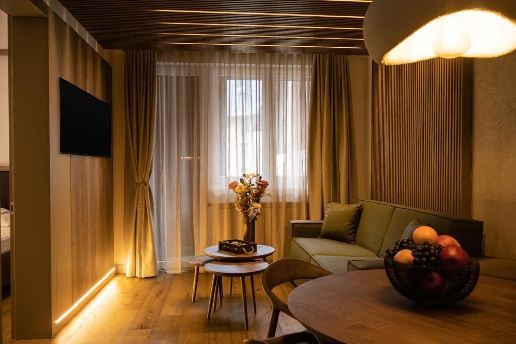 A modern living room with a wooden floor, a round dining table, a sofa, a TV mounted on the wall, and a window with sheer curtains.