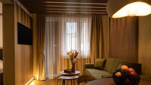 Una sala moderna con piso de madera, una mesa de comedor redonda, un sofá, un televisor montado en la pared y una ventana con cortinas traslúcidas.
