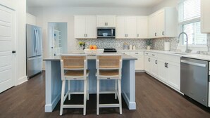 A modern kitchen with white cabinets, a central island with two wicker chairs, a refrigerator, a microwave, and a dishwasher. - Happy Ours, Mins to Park Circle, Dtwn, CHS Beach (North Charleston)