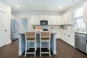 A modern kitchen with white cabinets, a central island with two wicker chairs, a refrigerator, a microwave, and a dishwasher. - Happy Ours, Mins to Park Circle, Dtwn, CHS Beach (North Charleston)