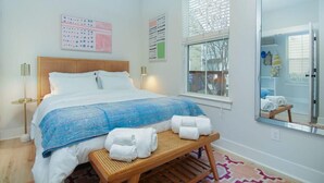 A neatly arranged bedroom with a bed, bedside lamps, a bedside table with towels, a window with blinds, and a mirror reflecting the room. - Happy Ours, Mins to Park Circle, Dtwn, CHS Beach (North Charleston)