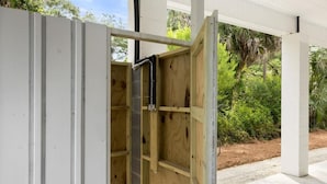 An open wooden structure with a black pipe, possibly a sauna or hot tub, set against a backdrop of greenery and a white building. - Jungle House-heated Pool-pet Friendly-luxury (Edisto Island)
