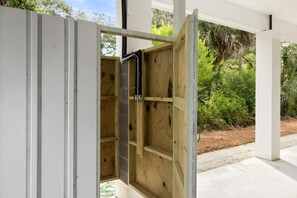 Una estructura de madera abierta con un tubo negro, posiblemente una sauna o bañera de hidromasaje, con un fondo de follaje y un edificio blanco.
