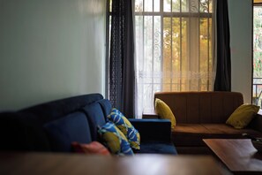 En stue med en blå sofa, en brun sofa med gule puder, samt et vindue med dekorative jerngitter og lette gardiner.