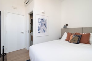 Um quarto com uma cama branca, uma pintura abstrata emoldurada, um abajur e uma cabeceira com travesseiros.