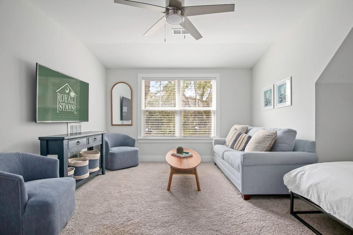 A modern, well-lit room with a ceiling fan, a large window with blinds, a grey sofa, a blue armchair, a wooden coffee table, and a bed with white bedding.