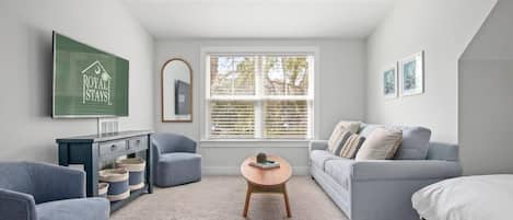 A modern, well-lit room with a ceiling fan, a large window with blinds, a grey sofa, a blue armchair, a wooden coffee table, and a bed with white bedding.