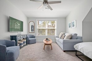 Een moderne, goed verlichte kamer met een plafondventilator, een groot raam met zonwering, een grijze bank, een blauwe fauteuil, een houten salontafel en een bed met witte beddengoed.