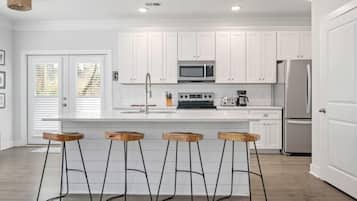 A modern kitchen with white cabinets, a stainless steel refrigerator, a microwave, and a coffee maker. There are four wooden stools with metal legs.