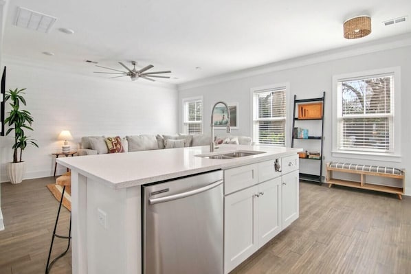 Una cocina moderna con muebles blancos, lavavajillas integrado y una isla grande con pileta. Hay ventanas con persianas, un ventilador de techo y un banco junto a la ventana.