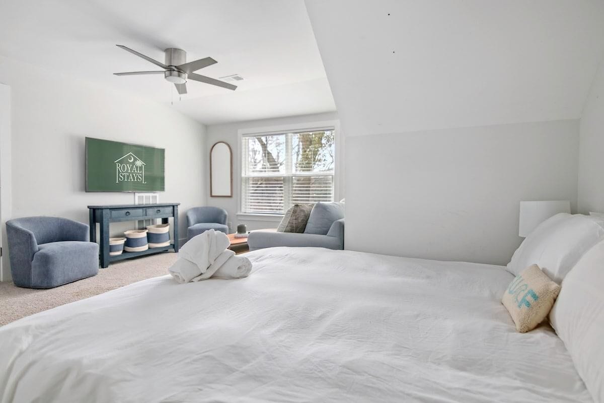 A bedroom with a large bed, a ceiling fan, a TV, and a window with blinds.