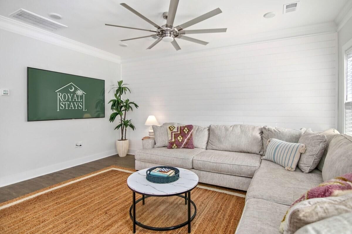 Uma sala de estar moderna com um sofá cinza, uma mesa de centro redonda, um ventilador de teto e uma TV na parede exibindo "ROYAL STAYS".