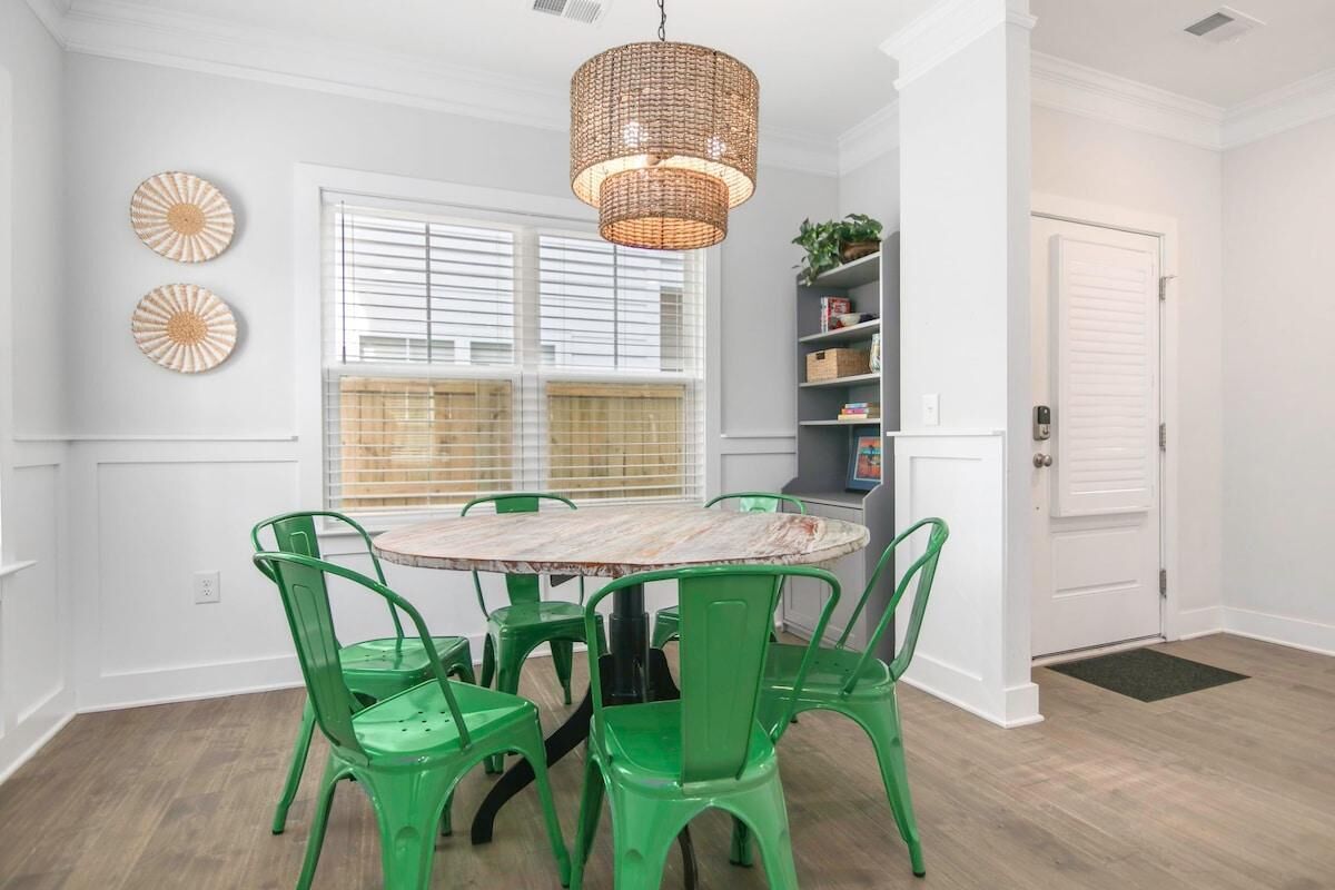 A dining area with a round table, green chairs, a wicker chandelier, and a window with blinds.