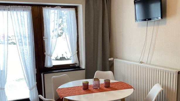 Un comedor con una mesa redonda, cuatro sillas, un televisor montado en la pared y una ventana con cortinas.