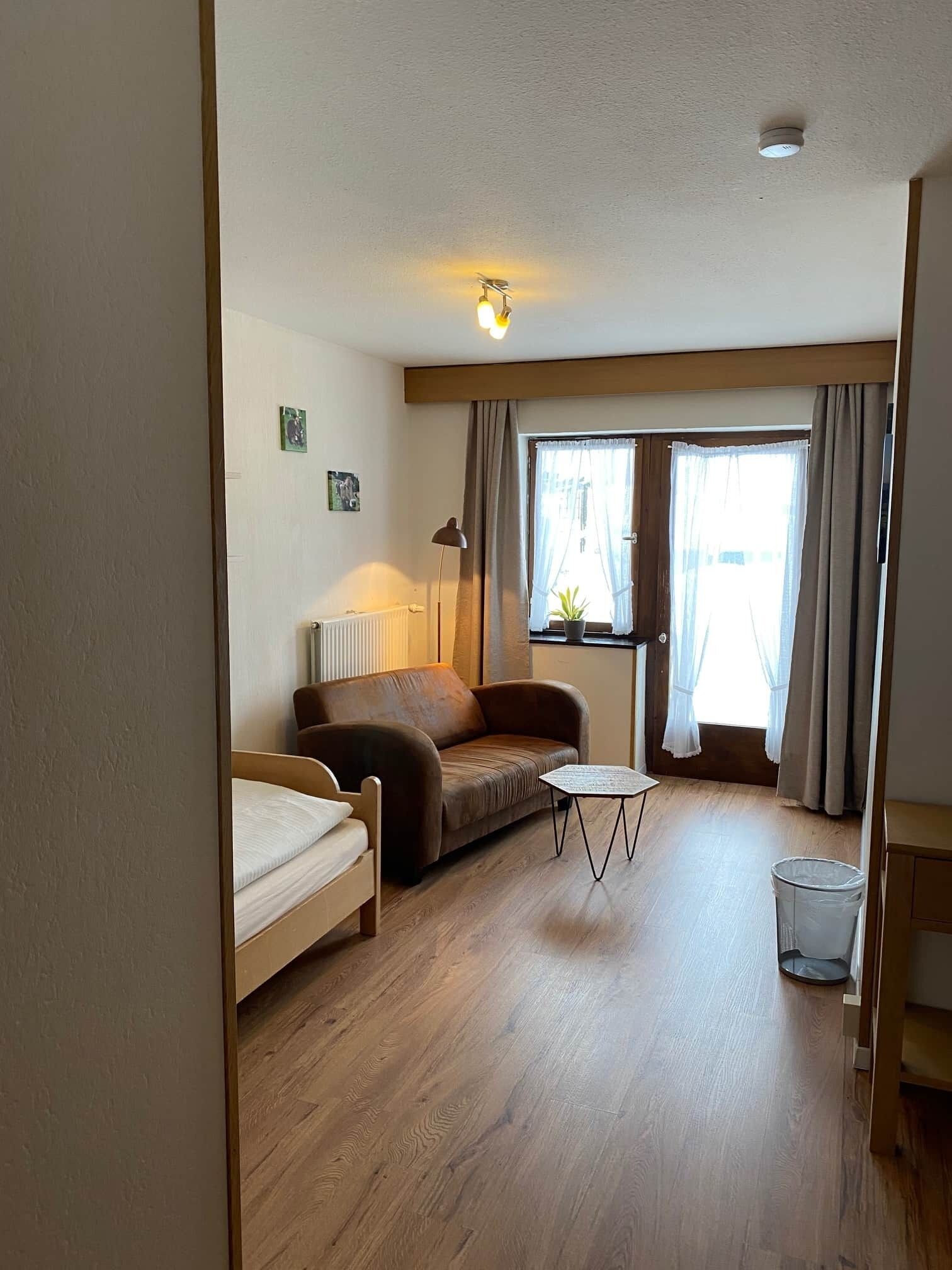 A furnished room with a bed, a sofa, a small table, a radiator, a lamp, and a window with curtains.