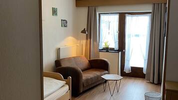 A furnished room with a bed, a sofa, a small table, a radiator, a lamp, and a window with curtains.