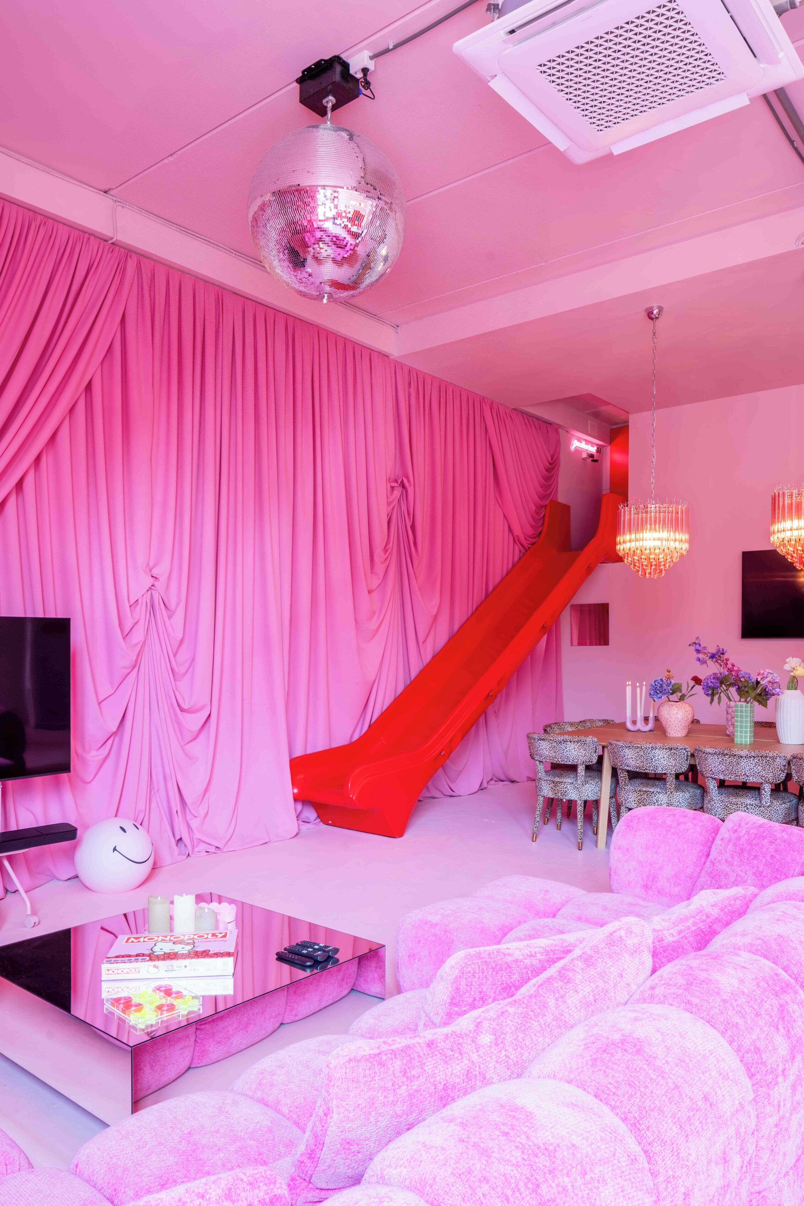 Una habitació amb cortines roses, una boleta de discoteca, un tobogan vermell, un joc de Monopoly i un sofà rosa.