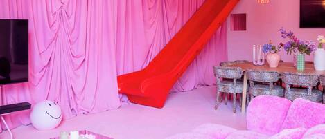 A room with pink curtains, a disco ball, a red slide, a Monopoly board game, and a pink sofa.