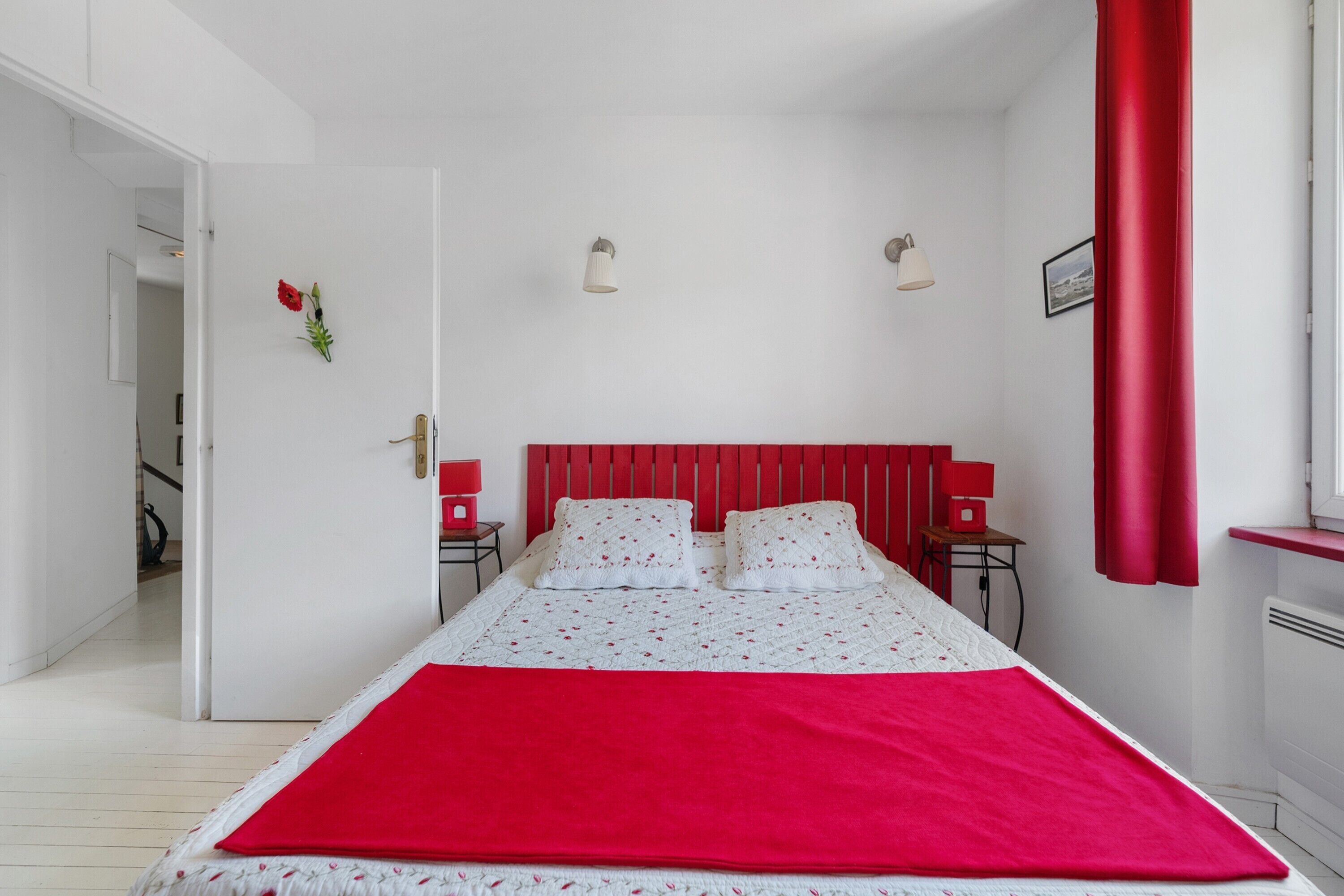 A bedroom with a bed, two bedside tables, a red headboard, and a window with red curtains.