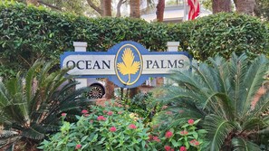 Un letrero con 'OCEAN PALMS' y el logotipo de una palmera, rodeado de exuberante follaje y plantas con flores.