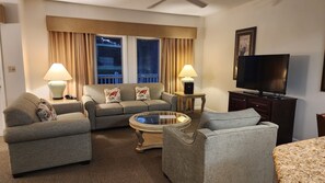 Ein Wohnzimmer mit einer Couch, zwei Sesseln, einem Couchtisch, einem Fernseher und einem Fenster mit Vorhängen.