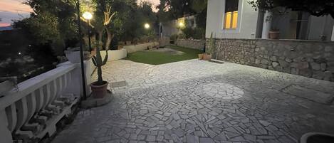 A stone-tiled patio with a lawn, surrounded by a white wall and a stone wall, with a cactus plant and potted plants.