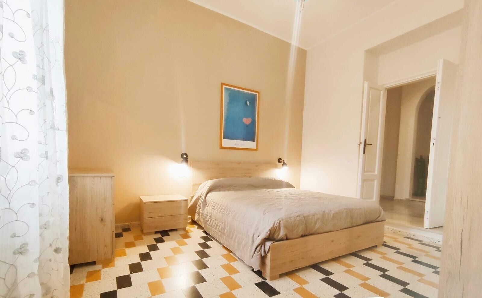 Ein Schlafzimmer mit einem Bett, Nachttischen, einer Wandleuchte, einer Tür und einem gemusterten Boden.