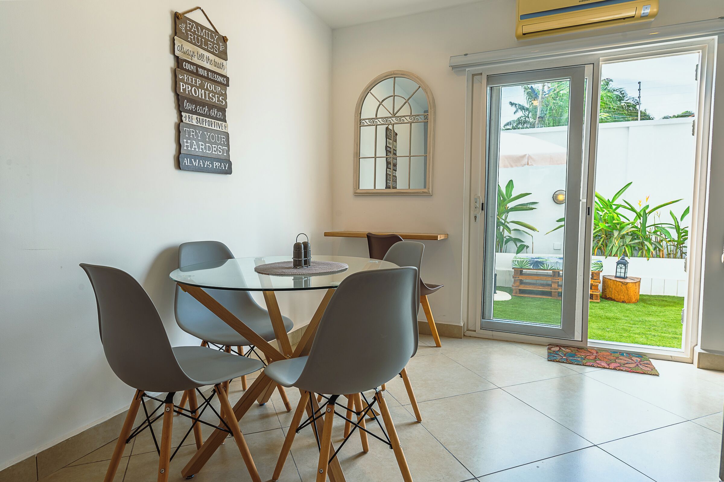 A dining area with a glass table, four chairs, a wall-mounted sign with family rules, a window with a view of a garden, and a sliding glass door.