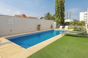 A swimming pool with a tiled edge, surrounded by artificial grass, potted plants, and a lounge area with a table and chairs. - Charming 4-bedroom townhome with AC, pool, and Wi-Fi in vibrant East Legon (Accra)