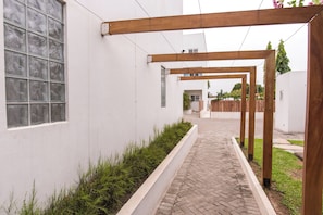 Un pasillo con una pérgola de madera, una pared blanca con ventana y un camino de ladrillo.