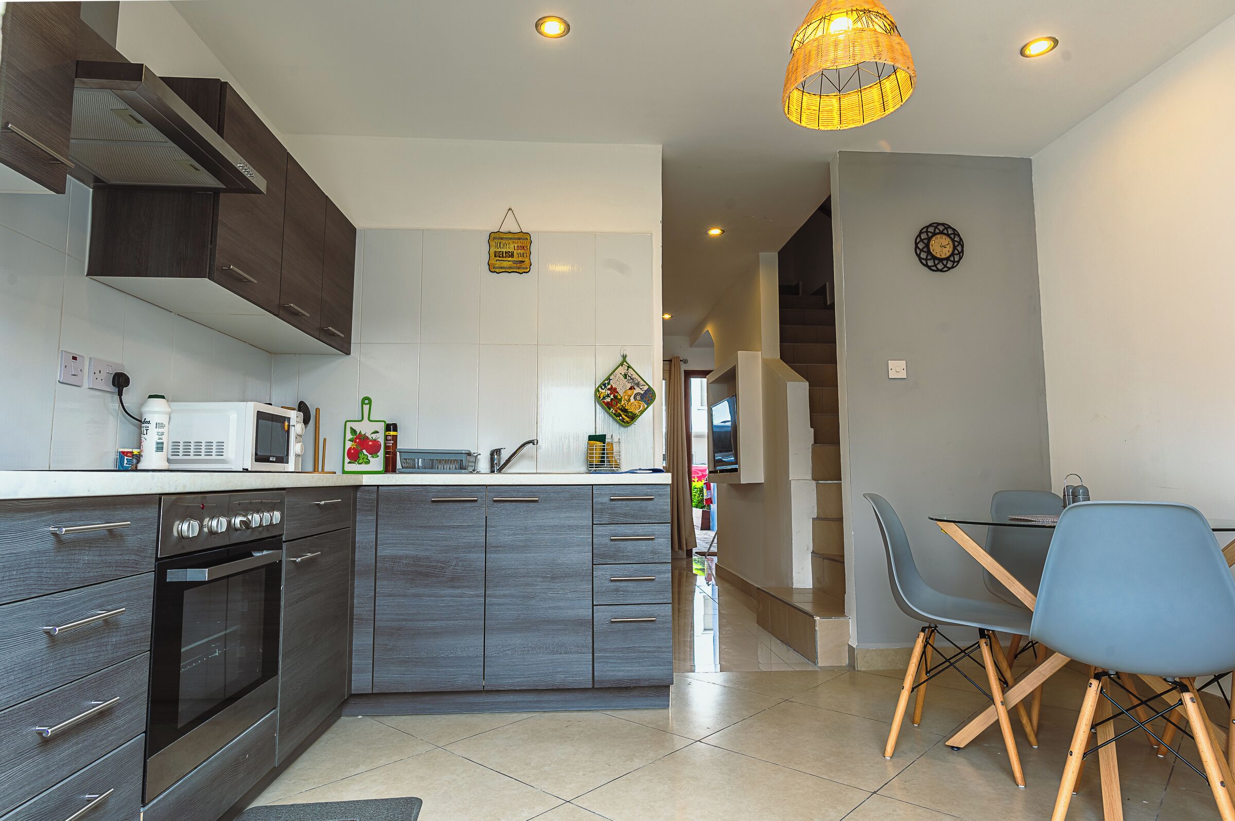 A modern kitchen with dark wood cabinets, a built-in oven, and a microwave. There is a dining area with a table and chairs, and a hanging light fixture.