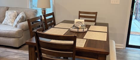A dining area with a wooden table, chairs, and a bench. There is a window with blinds, a lamp, and a framed picture on the wall.