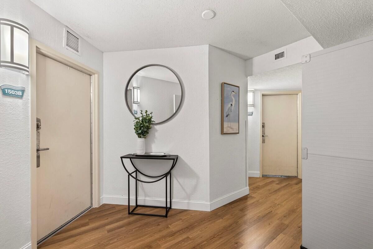 A hallway with wooden flooring, a round mirror on the wall, a small table with a plant, and a framed picture.