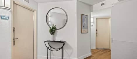 A hallway with wooden flooring, a round mirror on the wall, a small table with a plant, and a framed picture.