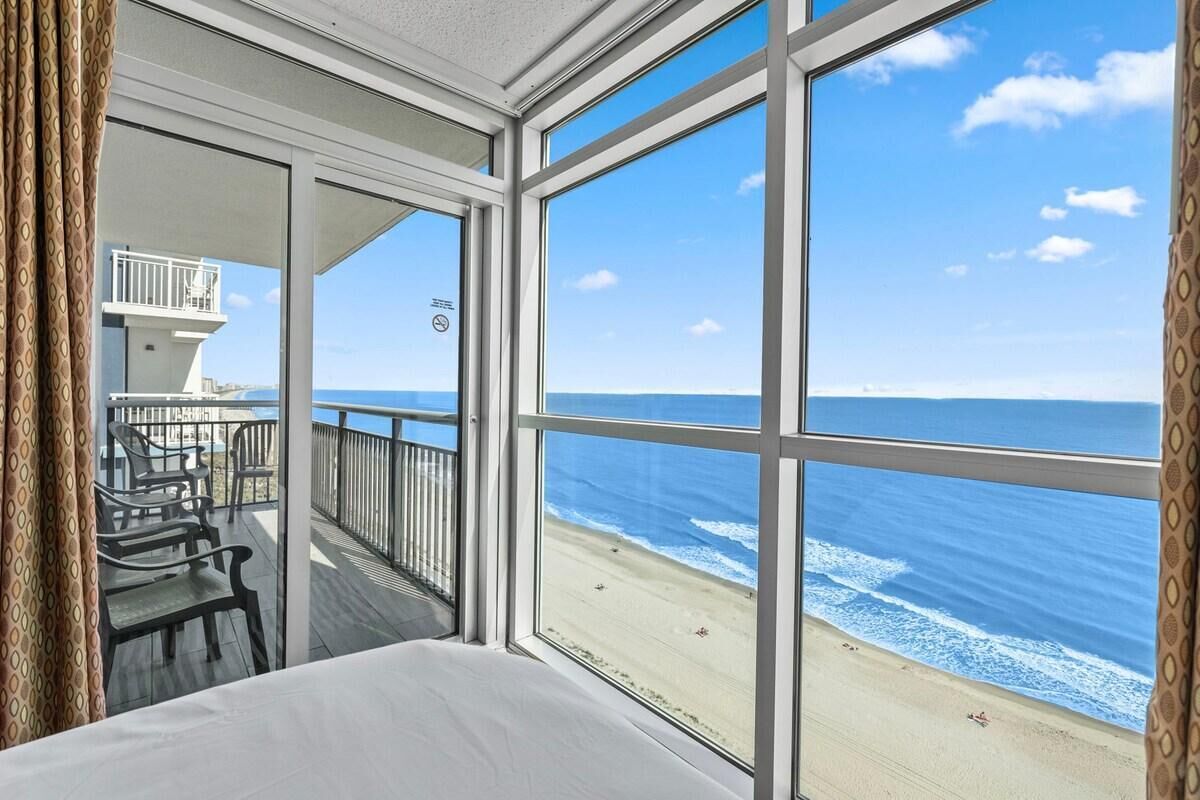 Une chambre dotée d’une grande fenêtre donnant sur une plage, d’un balcon avec des chaises et d’un lit.