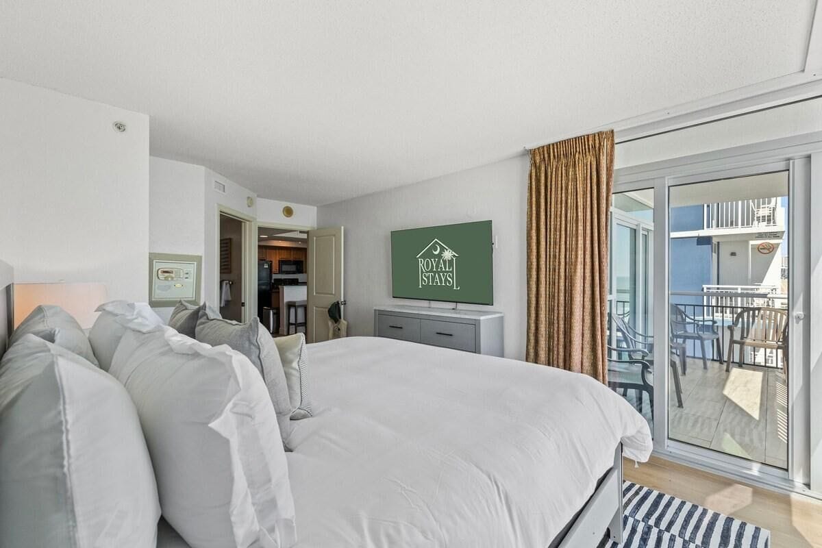 A bedroom with a large bed, a TV mounted on the wall, and a sliding glass door leading to an outdoor area with chairs.