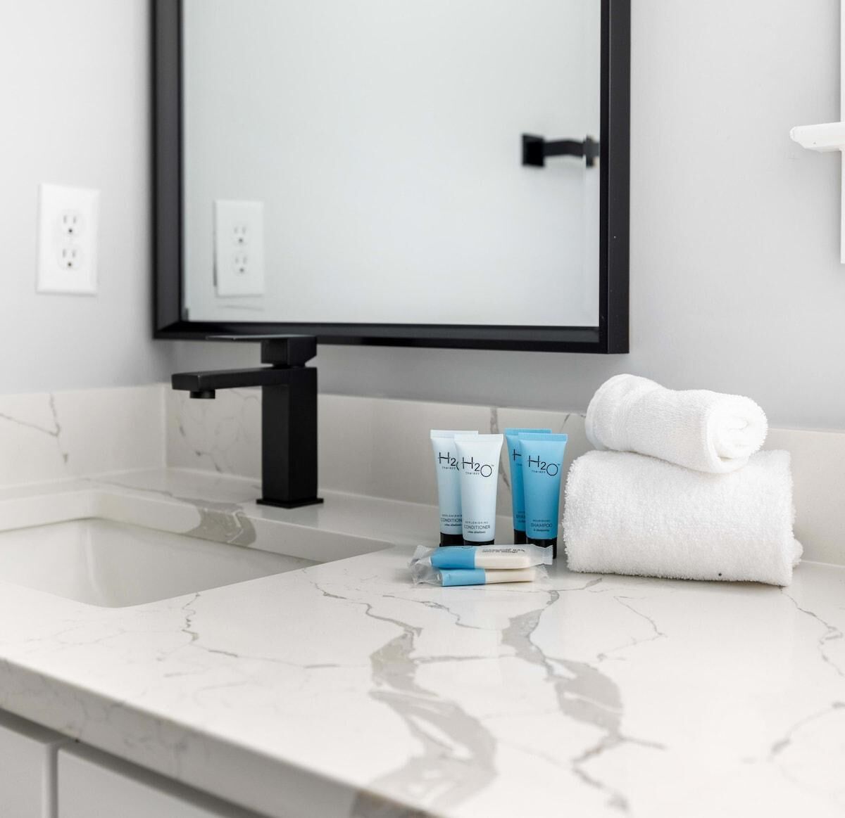 Ein Badezimmer mit einer weißen Marmorplatte, einem schwarzen Wasserhahn und Pflegeprodukten, darunter Flaschen von H2O Shampoo und Conditioner.