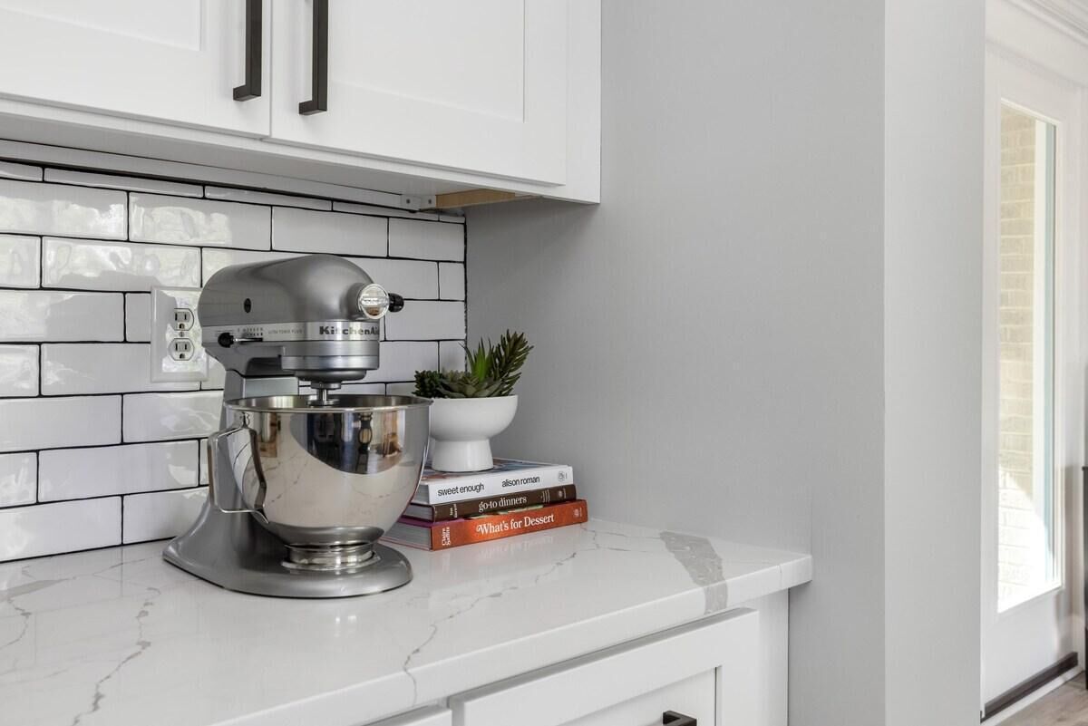 A kitchen with a stand mixer, a potted plant, and a stack of cookbooks on a white countertop.