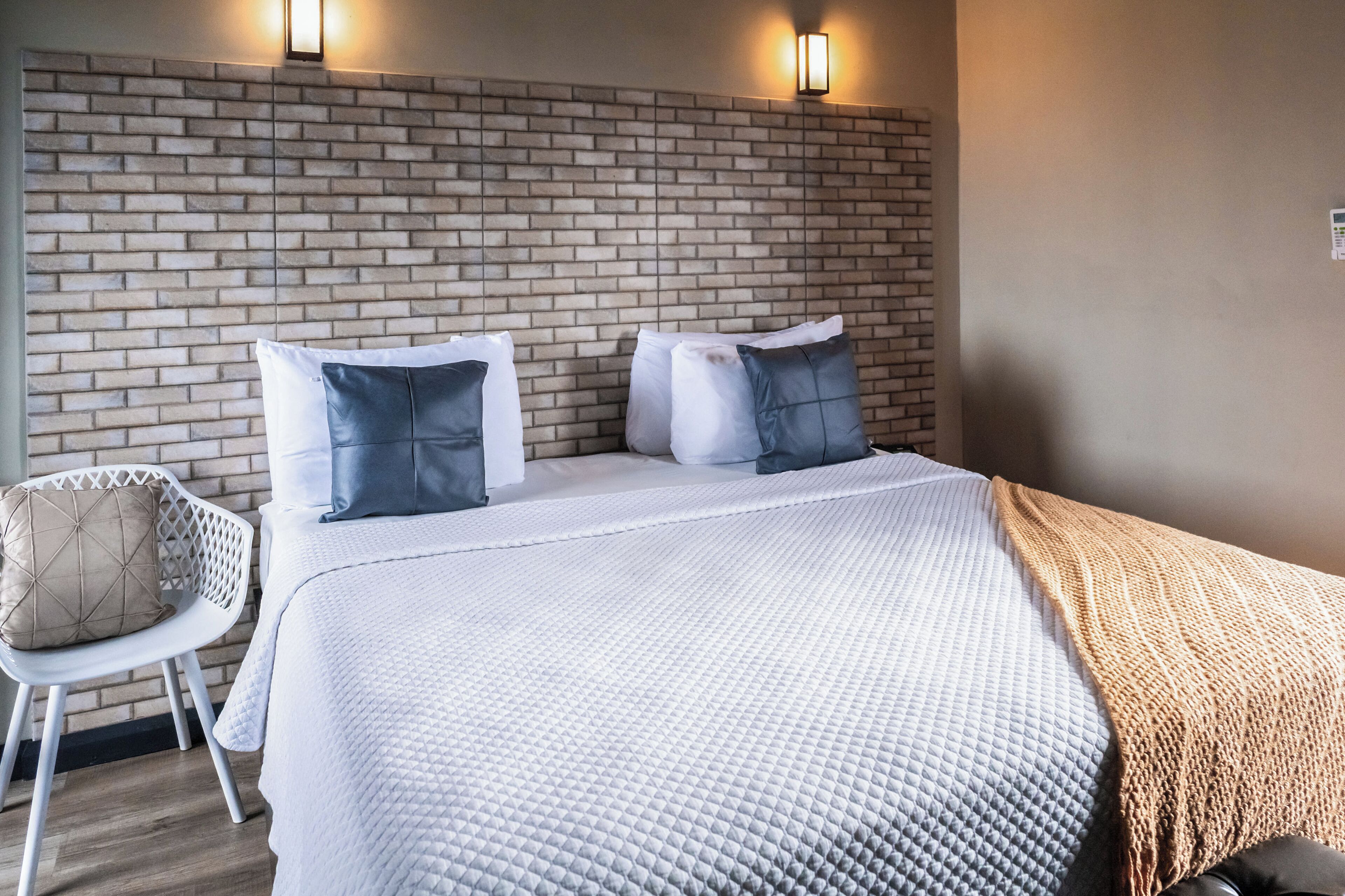 A hotel room with a brick-patterned headboard, a white bed with multiple pillows, a white chair, and a wooden floor.