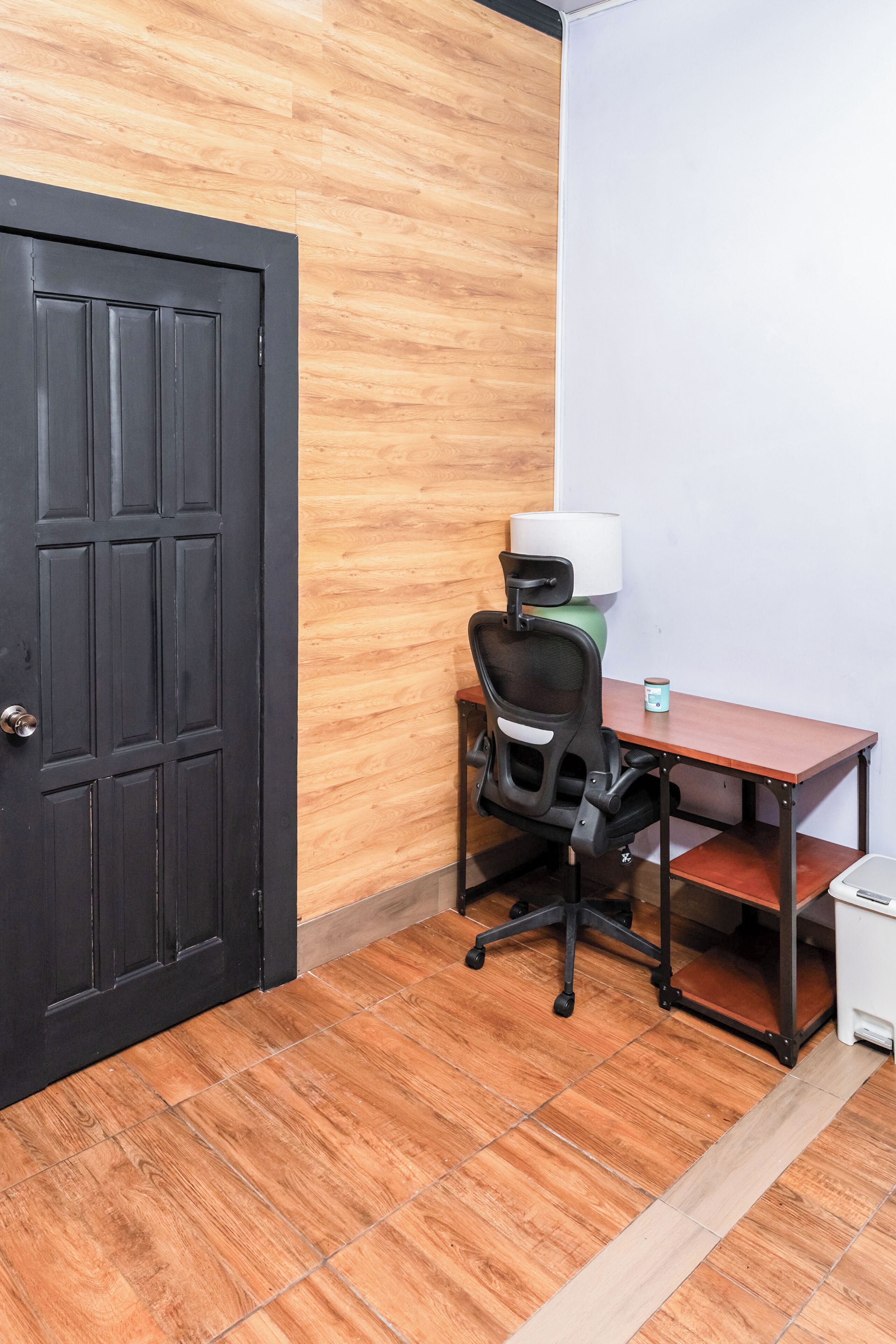 Une pièce avec un plancher en bois, une porte noire, un bureau avec une chaise, une lampe et une poubelle.